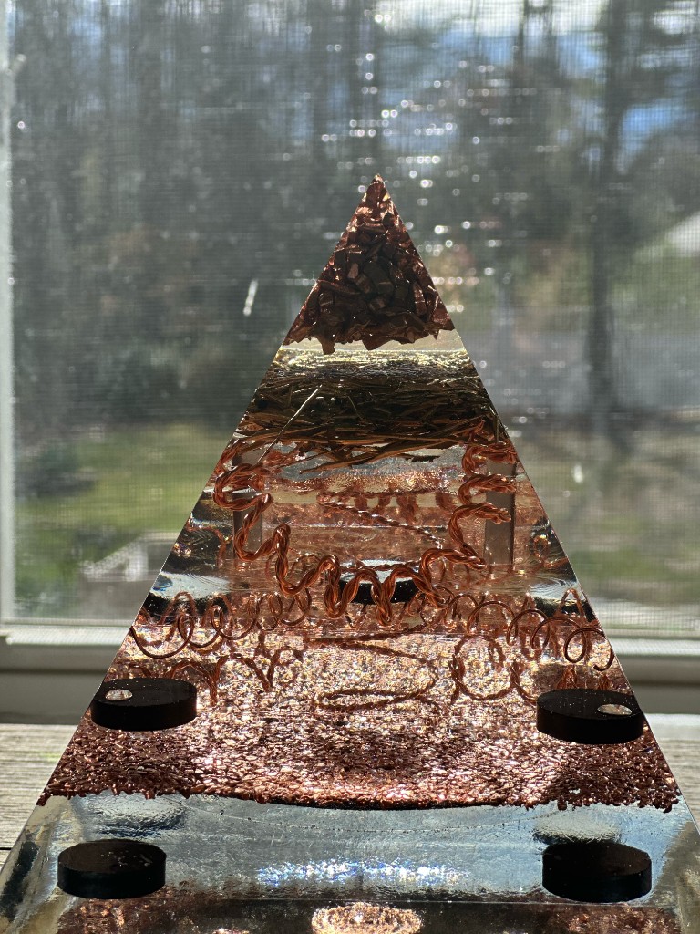 Large orgonite pyramid backlit against window showing copper coils and layered metals