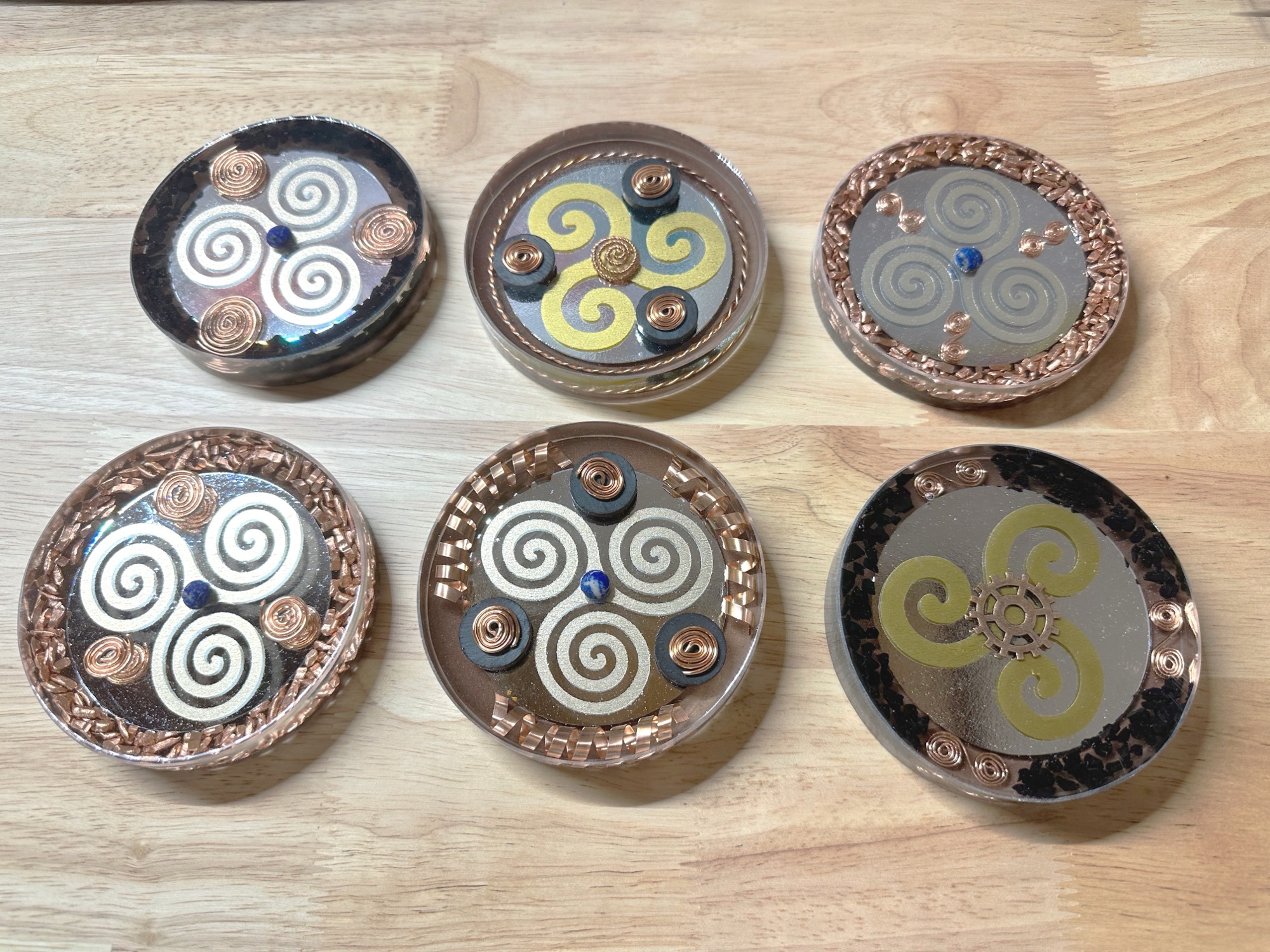 Six handmade Triskel Series orgone coasters arranged on dark wood, each with unique copper and sacred geometry detail